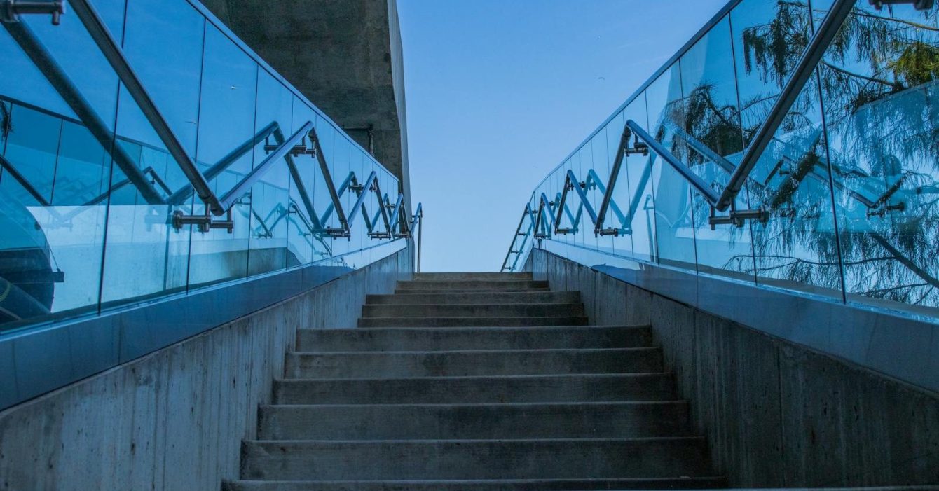 Photo by Jeremy Waterhouse Concrete stairs with glass railings reflecting blue sky, captured from a low angle.
