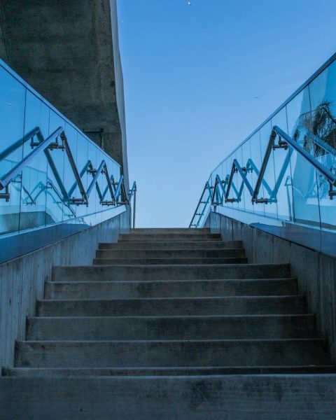Concrete stairs with glass railings reflecting blue sky, captured from a low angle.